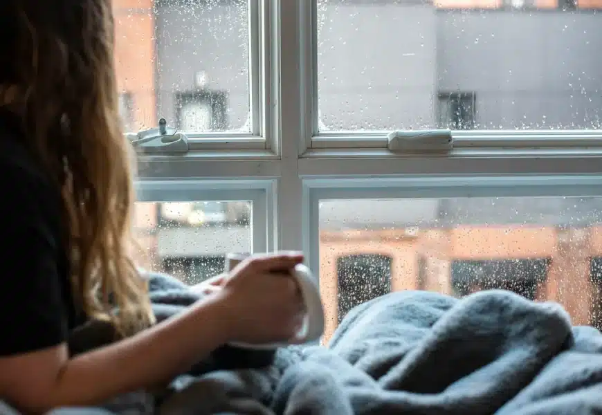 Jeune femme prenant son café devant sa fenêtre alors qu'il fait froid et pleut dehors