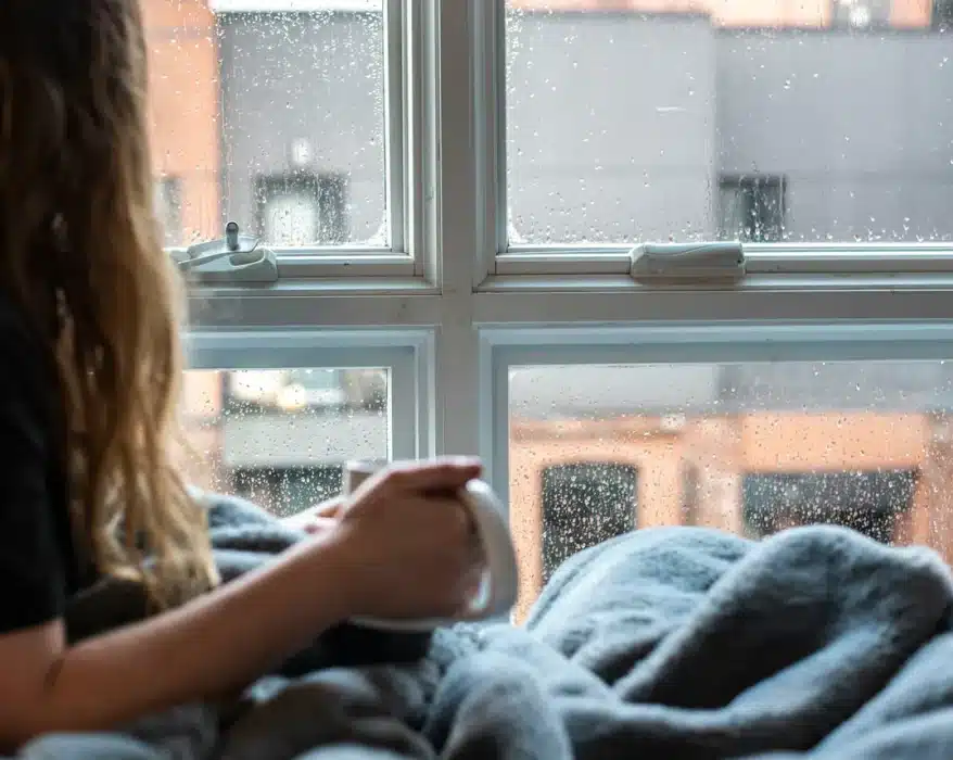 Jeune femme prenant son café devant sa fenêtre alors qu'il fait froid et pleut dehors